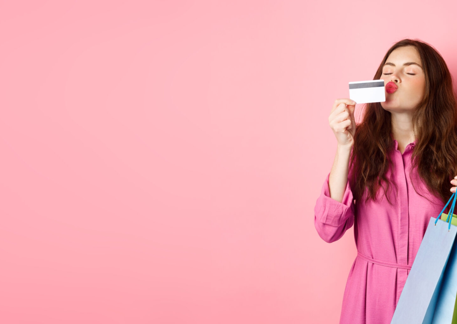 Portrait of excited happy shopaholic, woman holding shopping bags and kissing plastic credit card, smiling amazed, standing against pink background.